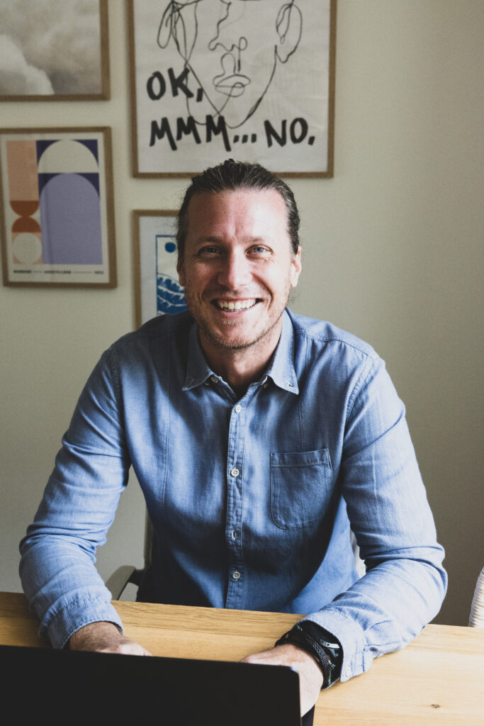 Christoph mit blauem Hemd sitzt vor seinem Laptop und grinst. Hinter ihm eine Wand mit unterschiedlichen Poster